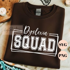 Può includere: Una felpa girocollo marrone scuro con testo bianco serigrafato che recita Dyslexia SQUAD in una combinazione di caratteri corsivi e in stampatello, racchiuso in una cornice rettangolare bianca. Il capo include un'etichetta sul collo GILDAN.