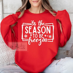 May include: Red sweatshirt with white text that reads 'Tis the season to be freezin' with snowflakes on either side of the text.