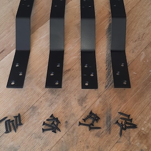 Może przedstawiać: Cztery czarne metalowe wsporniki i zestaw czarnych śrub ułożone na drewnianej powierzchni. Wsporniki są w kształcie litery L z wstępnie wywierconymi otworami do montażu. Śruby są rozrzucone w pobliżu.