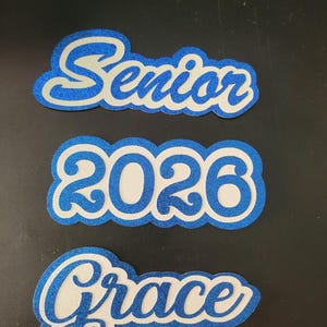 Pode incluir: Três recortes brilhantes azuis e brancos. O recorte superior diz "Senior" em uma fonte cursiva. O recorte do meio diz "2026". O recorte inferior diz "Grace" em uma fonte cursiva. Todos os recortes têm uma borda brilhante azul.