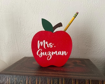 May include: A red wooden apple-shaped desk organizer with the name "Mrs. Guzman" in white script. A green leaf and brown stem are attached to the top. A yellow pencil with a pink eraser is inserted into the apple.