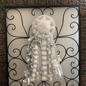 Puede incluir: Un adorno de homecoming mum en blanco y plata que presenta una gran roseta central con un centro de pompón blanco esponjoso, rodeado de cintas de satén en capas, detalles en plata metálica, pequeños lazos decorativos y tiras plateadas.