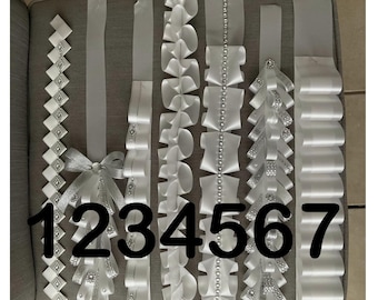 May include: Seven white satin ribbon homecoming mums or spirit garters labeled 1 through 7. Each features unique folding patterns, silver glitter accents, rhinestone embellishments, and pearl-like beads arranged in vertical decorative designs.