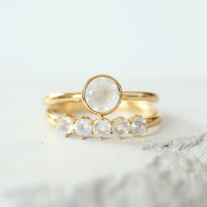 A set of two stackable rings placed on a concrete tray. The top ring is a moonstone solitaire ring in bezel setting. The bottom ring is a five stone ring that features small rainbow moonstone.