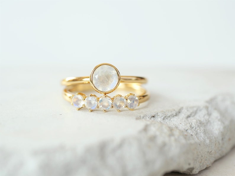 A set of two stackable rings placed on a concrete tray. The top ring is a moonstone solitaire ring in bezel setting. The bottom ring is a five stone ring that features small rainbow moonstone.