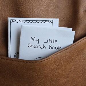 Könnte beinhalten: Ein kleines weißes Büchlein mit dem Titel "My Little Church Book" steckt in einer braunen Ledertasche. Das Büchlein hat einen dekorativen, wellenförmigen Rand. Die Tasche ist Teil eines größeren braunen Lederartikels.