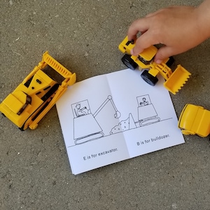 May include: A yellow toy excavator and a yellow toy bulldozer are shown next to a children's book. The book has black and white illustrations of an excavator and a bulldozer. The text on the book reads "E is for excavator" and "B is for bulldozer".