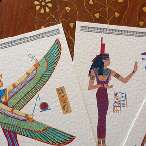 May include: Two cards with illustrations of Egyptian goddesses. The first card features a winged goddess with a blue and green headdress and a golden ankh symbol. The second card features a goddess with a blue headdress and a red and purple dress. Both cards have a white background with a decorative border.
