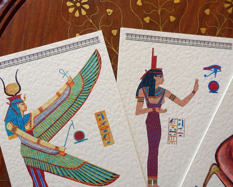 May include: Two cards with illustrations of Egyptian goddesses. The first card features a winged goddess with a blue and green headdress and a golden ankh symbol. The second card features a goddess with a blue headdress and a red and purple dress. Both cards have a white background with a decorative border.