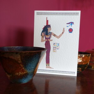 May include: A white card with a colorful illustration of an Egyptian goddess with a red and blue dress, holding her hand up to a blue eye symbol. The card is sitting on a wooden table with two brown and blue ceramic bowls.
