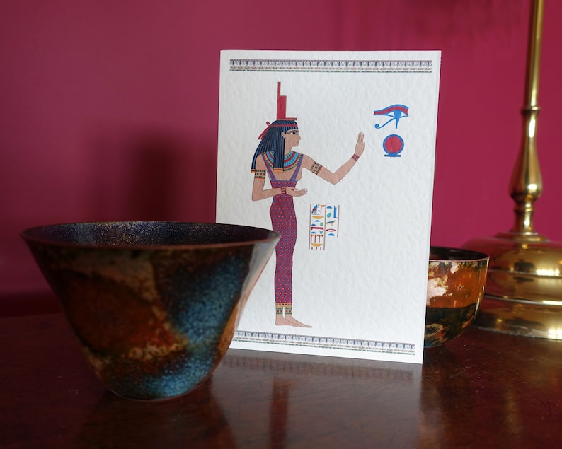 May include: A white card with a colorful illustration of an Egyptian goddess with a red and blue dress, holding her hand up to a blue eye symbol. The card is sitting on a wooden table with two brown and blue ceramic bowls.