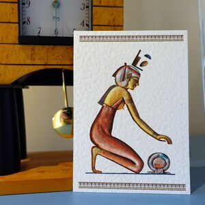 May include: A white card with a gold and red illustration of an Egyptian woman kneeling and holding a round object. The woman is wearing a gold crown and a red dress. The card has a gold border.