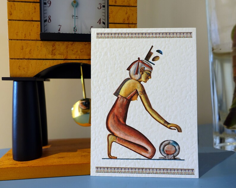 May include: A white card with a gold and red illustration of an Egyptian woman kneeling and holding a round object. The woman is wearing a gold crown and a red dress. The card has a gold border.