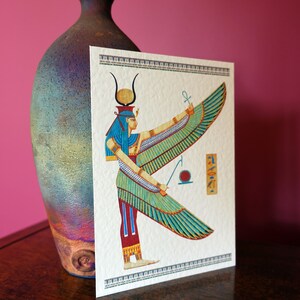 May include: A colorful illustration of the Egyptian goddess Isis with outstretched wings. She is depicted with a blue headdress and a golden crown. The background is white with a decorative border.