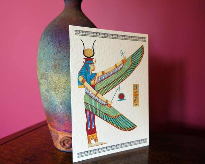 May include: A colorful illustration of the Egyptian goddess Isis with outstretched wings. She is depicted with a blue headdress and a golden crown. The background is white with a decorative border.