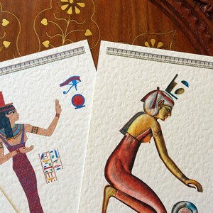 May include: Two vintage-style illustrations of Egyptian women. The woman on the left is wearing a blue and red dress and a headdress with a blue eye symbol. The woman on the right is wearing a red dress and a headdress with a blue and white design. Both illustrations are on a textured white background.