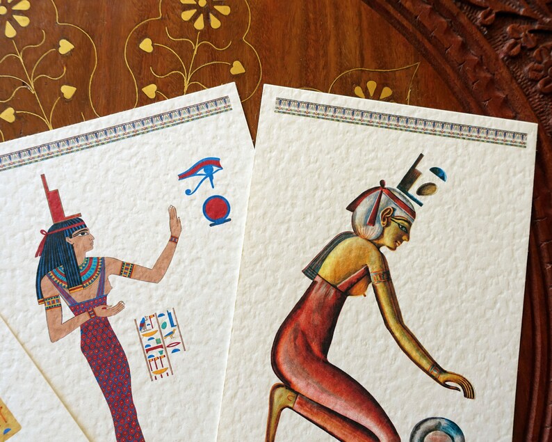 May include: Two vintage-style illustrations of Egyptian women. The woman on the left is wearing a blue and red dress and a headdress with a blue eye symbol. The woman on the right is wearing a red dress and a headdress with a blue and white design. Both illustrations are on a textured white background.