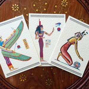 May include: Three vintage postcards featuring illustrations of Egyptian goddesses. The first postcard depicts a winged goddess with a blue headdress and a red and green dress. The second postcard shows a goddess with a blue headdress and a red dress, holding a staff. The third postcard features a goddess with a red dress and a blue headdress, kneeling and holding a ball.