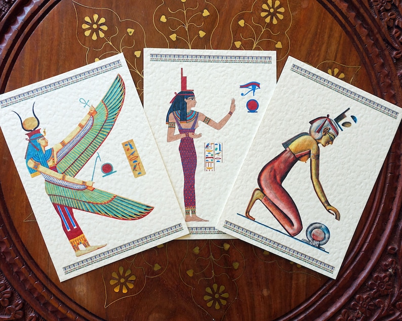 May include: Three vintage postcards featuring illustrations of Egyptian goddesses. The first postcard depicts a winged goddess with a blue headdress and a red and green dress. The second postcard shows a goddess with a blue headdress and a red dress, holding a staff. The third postcard features a goddess with a red dress and a blue headdress, kneeling and holding a ball.