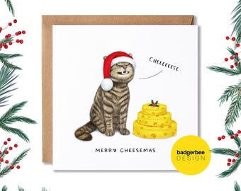 May include: A Christmas card featuring a cartoon cat wearing a Santa hat and saying "Cheeeese". The cat is sitting next to a stack of cheese shaped like a Christmas cake. The card reads "Merry Cheesemas" and has the text "badgerbee design" in the bottom right corner.