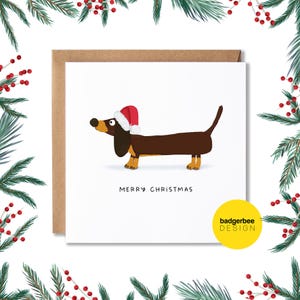 Pu&ograve; includere: Un biglietto di auguri di Natale con un bassotto di cartone animato che indossa un cappello di Babbo Natale. Il biglietto ha il testo "Buon Natale" e un piccolo logo che dice "badgerbee DESIGN".