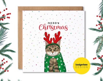 May include: A Christmas card featuring a cat wearing a green sweater and reindeer antlers. The card has a white background with colorful confetti and the text "Merry Christmas".