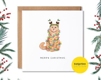 May include: A Christmas card featuring a cartoon orange cat wearing reindeer antlers and wrapped in colorful Christmas lights. The card reads "Merry Christmas" and has the text "badgerbee DESIGN" in the bottom right corner.