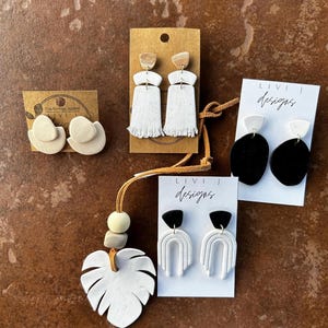 May include: Assortment of handmade polymer clay earrings in various styles and colors, including white, black, and brown. The earrings are displayed on cards with the text "Livi J Designs".