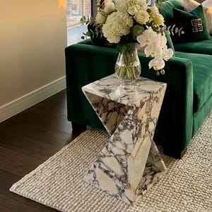 Pode incluir: Uma sala de estar luxuosa com um sofá de veludo verde escuro, uma mesa lateral de mármore e uma obra de arte abstrata emoldurada. A sala é preenchida com luz natural e inclui um vaso com flores brancas e uma planta em vaso, criando uma atmosfera sofisticada e convidativa.