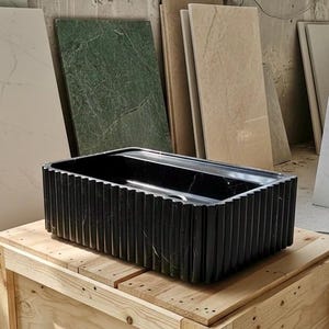 Pode incluir: Uma pia retangular preta de pedra com detalhes verticais canelados. A pia é exibida em uma caixa de madeira, com várias placas de pedra ao fundo. As dimensões da pia são de aproximadamente 76 cm de comprimento, 38 cm de largura e 15 cm de profundidade.