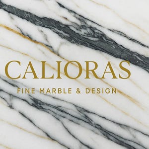May include: White marble surface with gold and gray veining. The text "CALIORAS FINE MARBLE & DESIGN" is displayed in gold lettering. The marble has a diagonal pattern.