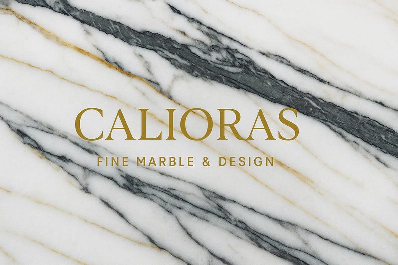 May include: White marble surface with gold and gray veining. The text "CALIORAS FINE MARBLE & DESIGN" is displayed in gold lettering. The marble has a diagonal pattern.