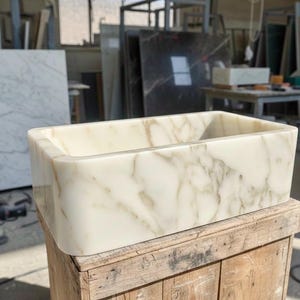 Op de afbeelding: Rechthoekige marmeren wastafel in crème- en beige tinten met een geaderd patroon. De wastafel staat op een houten krat in een werkplaats. De binnenkant van de wastafel is een glad, open bekken.