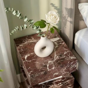 K&ouml;nnte beinhalten: Ein Schlafzimmer mit einem Nachttisch aus rotem Marmor. Auf dem Tisch steht eine wei&szlig;e Vase mit einer wei&szlig;en Rose und Gr&uuml;n. Der Nachttisch besteht aus zwei gestapelten rechteckigen Bl&ouml;cken. Das Bett hat ein graues Kopfteil.