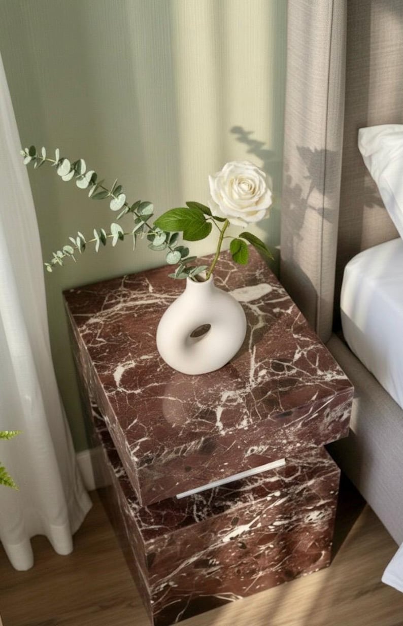 K&ouml;nnte beinhalten: Ein Schlafzimmer mit einem Nachttisch aus rotem Marmor. Auf dem Tisch steht eine wei&szlig;e Vase mit einer wei&szlig;en Rose und Gr&uuml;n. Der Nachttisch besteht aus zwei gestapelten rechteckigen Bl&ouml;cken. Das Bett hat ein graues Kopfteil.