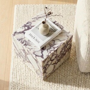 May include: A square marble side table with a purple and white veined pattern. A small beige vase and a book with the word "ROCKNIE" are on top. The table sits on a white textured rug, next to a beige armchair.