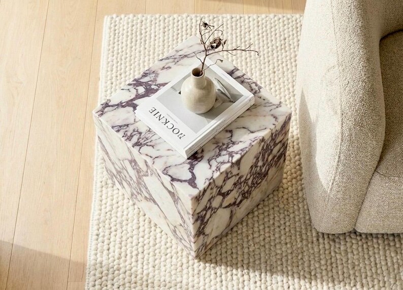 May include: A square marble side table with a purple and white veined pattern. A small beige vase and a book with the word "ROCKNIE" are on top. The table sits on a white textured rug, next to a beige armchair.