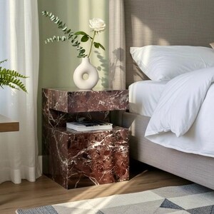 K&ouml;nnte beinhalten: Ein Schlafzimmer-Interieur mit einem Nachttisch in roter Marmoroptik. Eine wei&szlig;e Vase mit einer wei&szlig;en Rose und Bl&auml;ttern steht darauf. Der Tisch steht neben einem Bett mit neutralem Kopfteil und wei&szlig;er Bettw&auml;sche. Ein geometrischer Teppich liegt auf dem Boden.