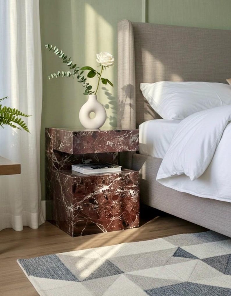 K&ouml;nnte beinhalten: Ein Schlafzimmer-Interieur mit einem Nachttisch in roter Marmoroptik. Eine wei&szlig;e Vase mit einer wei&szlig;en Rose und Bl&auml;ttern steht darauf. Der Tisch steht neben einem Bett mit neutralem Kopfteil und wei&szlig;er Bettw&auml;sche. Ein geometrischer Teppich liegt auf dem Boden.