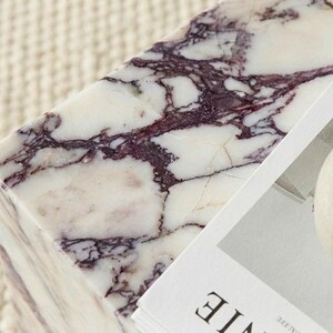 May include: A rectangular marble block with a white base and striking purple veining. The stone surface is next to a stack of white paper and a small, beige vase. The overall aesthetic is minimalist and elegant.