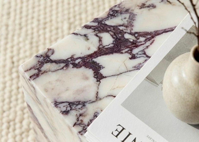 May include: A rectangular marble block with a white base and striking purple veining. The stone surface is next to a stack of white paper and a small, beige vase. The overall aesthetic is minimalist and elegant.