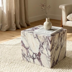 May include: A square marble side table with a white and purple veined pattern. A small white vase with a sprig of dried flowers and a book with the word "MOCKNIE" are on top. The table sits on a cream-colored rug.