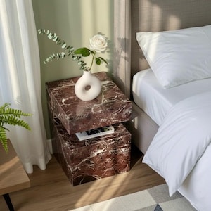 Könnte beinhalten: Ein modernes Schlafzimmer mit einem gestapelten Nachttisch aus Marmor. Der Nachttisch ist rotbraun mit weißen Adern und trägt eine weiße Vase mit einer weißen Rose und Grün. Ein Bett mit weißer Bettwäsche und ein geometrischer Teppich sind ebenfalls zu sehen.