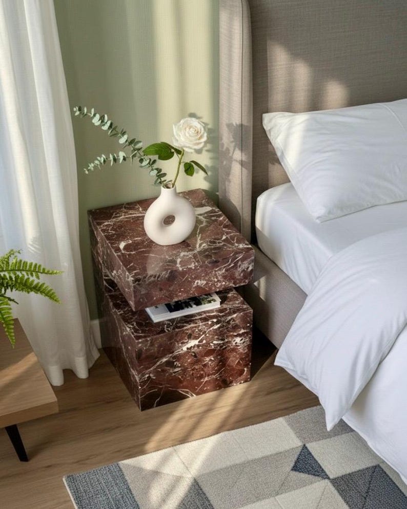 K&ouml;nnte beinhalten: Ein modernes Schlafzimmer mit einem gestapelten Nachttisch aus Marmor. Der Nachttisch ist rotbraun mit wei&szlig;en Adern und tr&auml;gt eine wei&szlig;e Vase mit einer wei&szlig;en Rose und Gr&uuml;n. Ein Bett mit wei&szlig;er Bettw&auml;sche und ein geometrischer Teppich sind ebenfalls zu sehen.