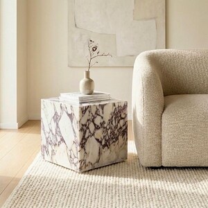 May include: A modern living room scene featuring a square marble side table with purple veining, topped with books and a small vase. A beige boucle sofa sits nearby on a cream-colored rug, with a large abstract painting on the wall.