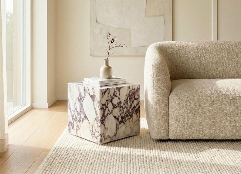 May include: A modern living room scene featuring a square marble side table with purple veining, topped with books and a small vase. A beige boucle sofa sits nearby on a cream-colored rug, with a large abstract painting on the wall.