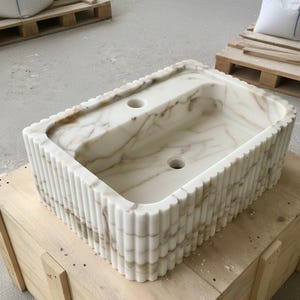 May include: Rectangular white marble sink with a fluted exterior design. The sink has two tap holes and a basin with brown veining. The sink is sitting on a wooden crate.