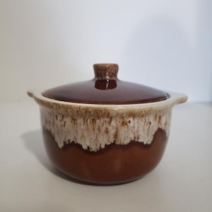 May include: A brown ceramic casserole dish with a lid. The dish has a rounded shape with a white and brown speckled rim. The lid is a darker shade of brown with a small handle. The dish is designed for cooking and serving.