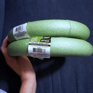 May include: Two circular green floral foam rings for crafting projects. Each ring features a white product label with the text "CRAFT ARTISAN FLORAL FOAM" and "9.6 in (24 cm)" printed on the surface, along with a barcode and safety warnings.