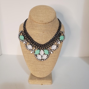May include: A black statement necklace with a multi-strand chain design. The necklace features large, oval-shaped, mint green and white gemstones, accented with smaller clear crystals. The necklace is displayed on a tan jewelry bust.